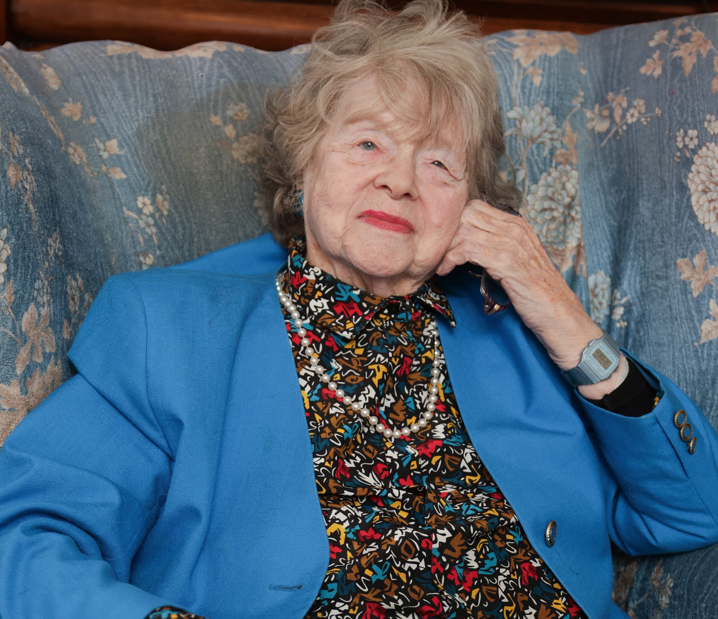 Elderly woman in a blue jacket sits on a floral-patterned couch, resting her head on her hand. - Home Instead