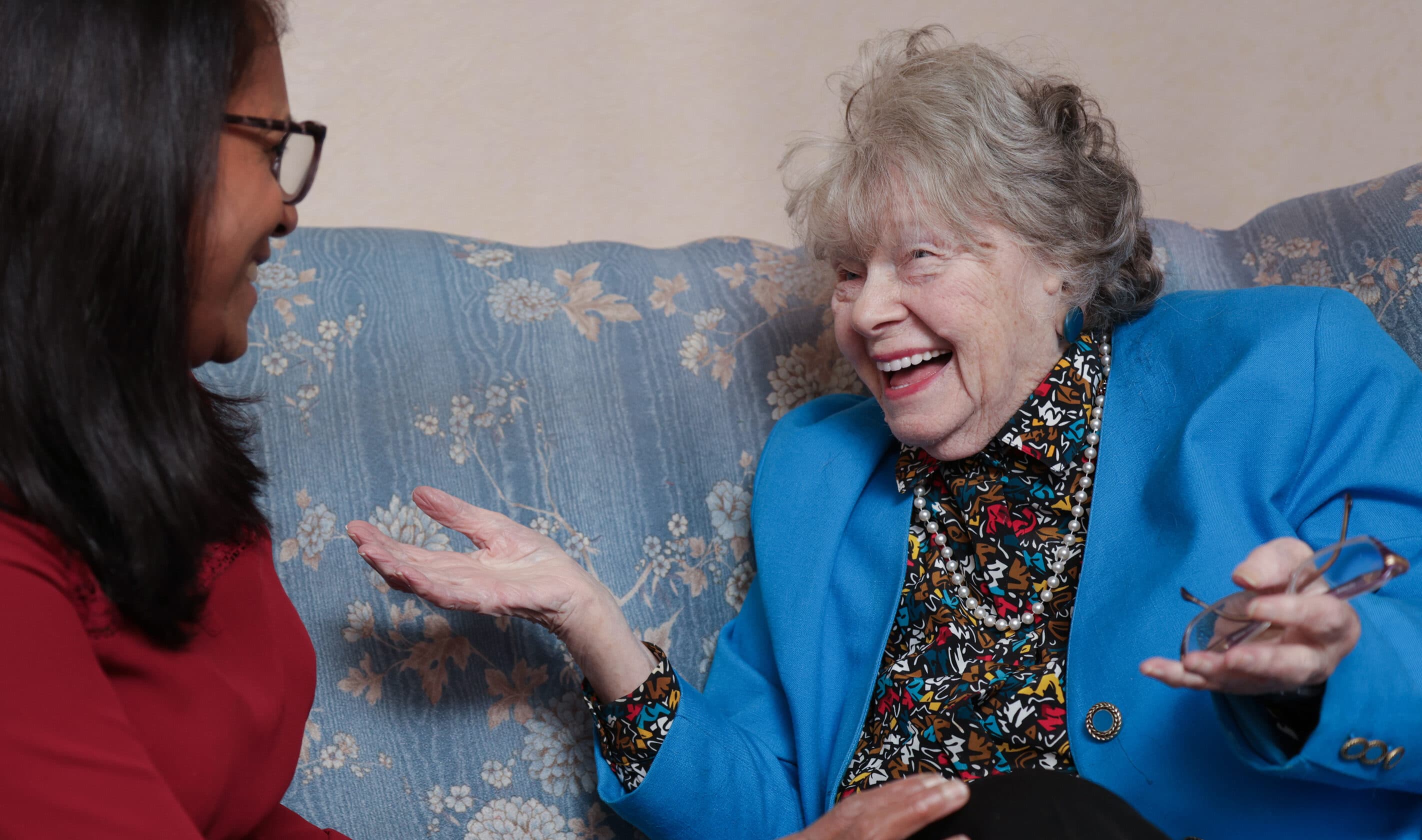 An elderly woman laughing and chatting with a younger woman on a blue patterned sofa. - Home Instead