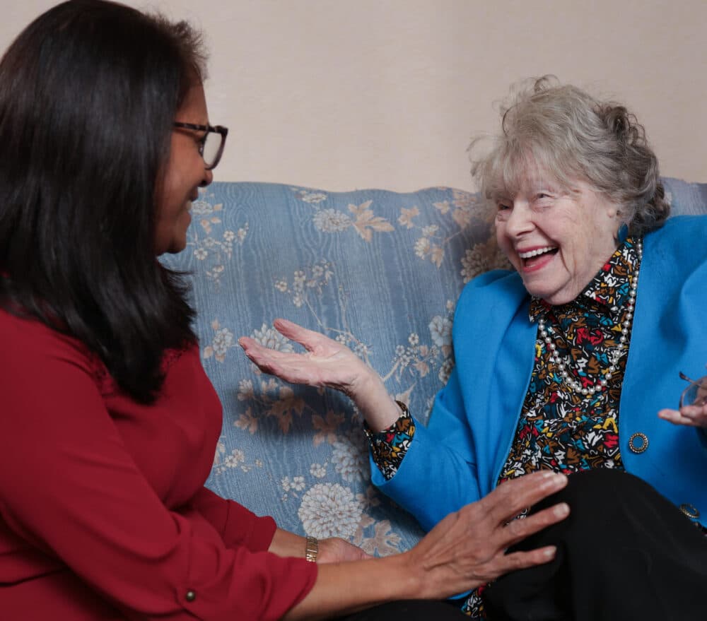 Two women sit on a couch, smiling and laughing while having an animated conversation. - Home Instead