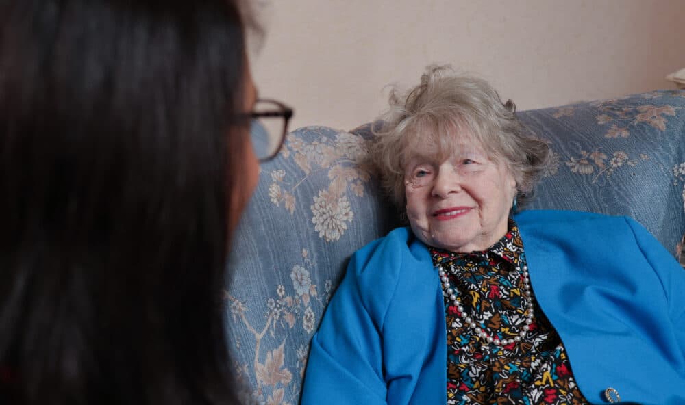 An elderly woman in a blue jacket smiles while talking to someone on a floral-patterned sofa. - Home Instead