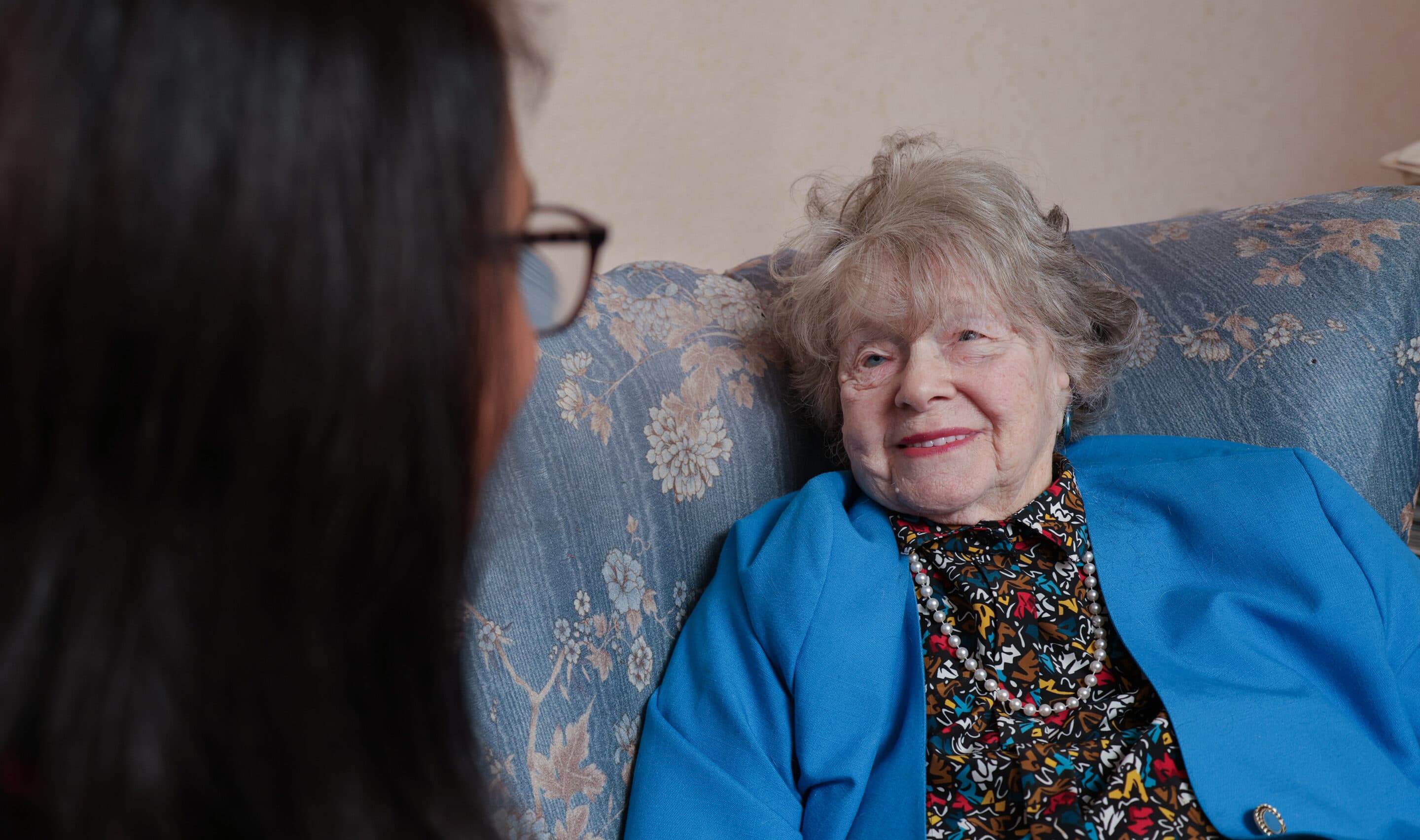 An elderly woman in a blue jacket smiles while talking to someone on a floral-patterned sofa. - Home Instead