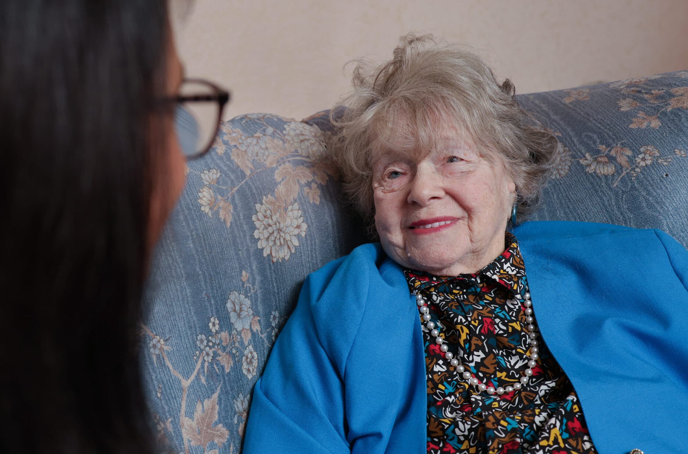Elderly woman in a blue jacket smiles while sitting on a patterned couch, speaking to another person. - Home Instead