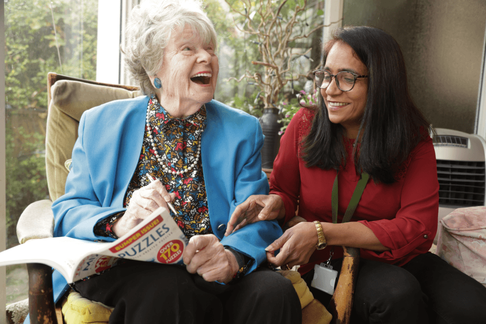 An elderly woman and a younger woman laugh together while doing a puzzle book in a bright room. - Home Instead