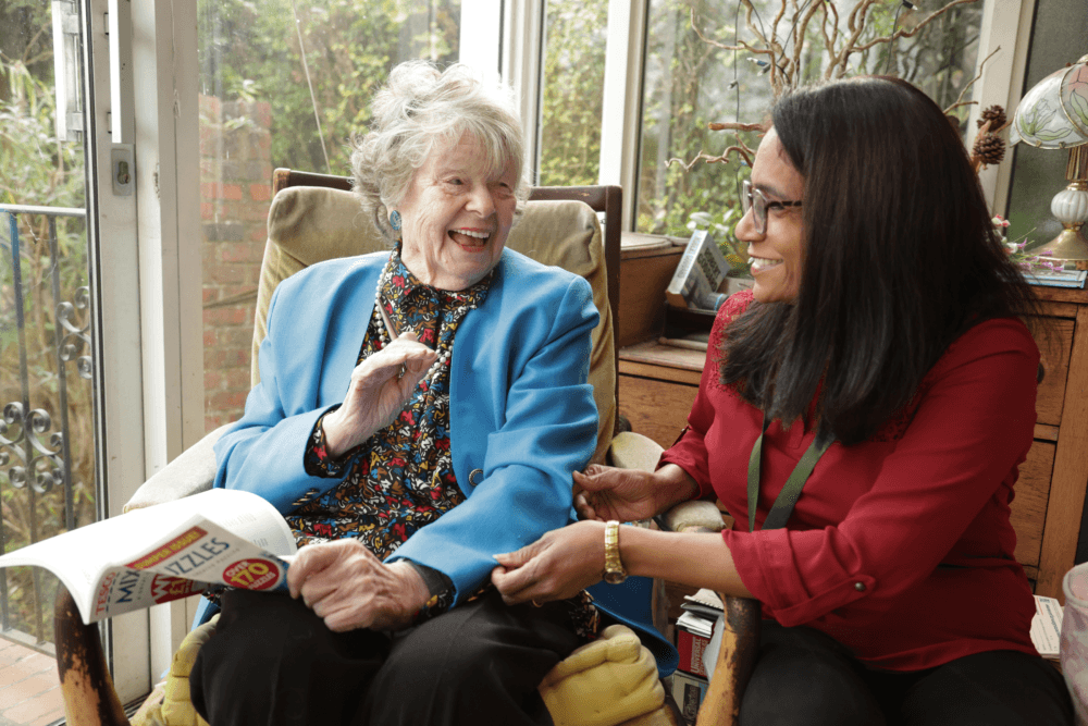 An elderly woman and a younger woman smile and laugh together while sitting indoors by a window. - Home Instead