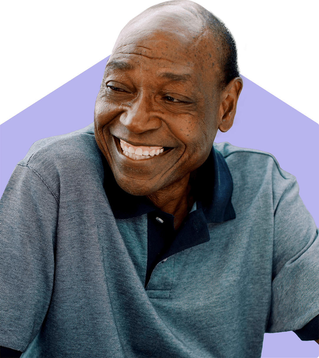 Smiling older man in a gray polo shirt, seated in front of a purple geometric background. - Home Instead