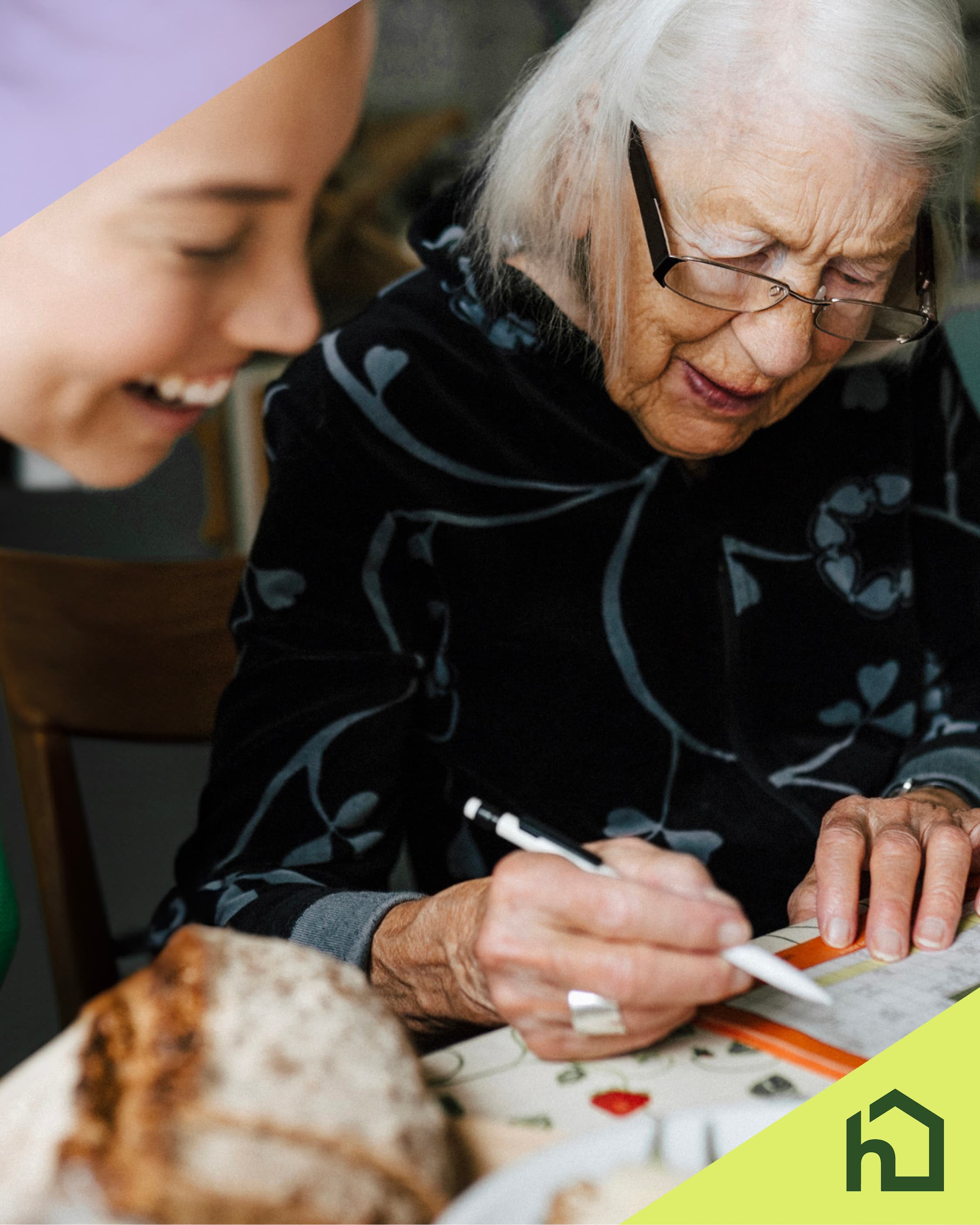 Elderly woman writing with a pen while a smiling caregiver sits beside her at a table. - Home Instead