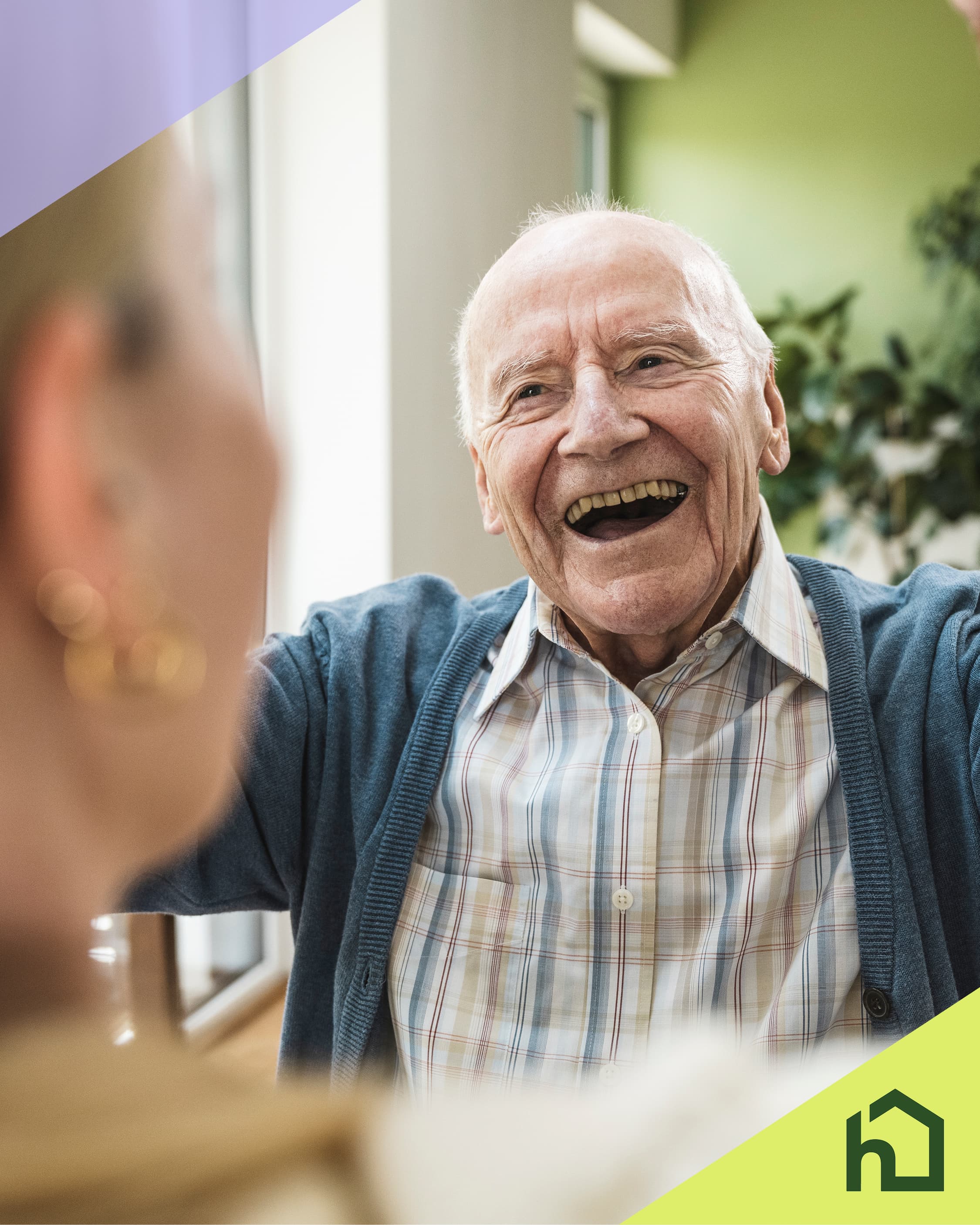 An elderly man smiling and laughing indoors, facing a blurred person, with a green logo in the corner. - Home Instead