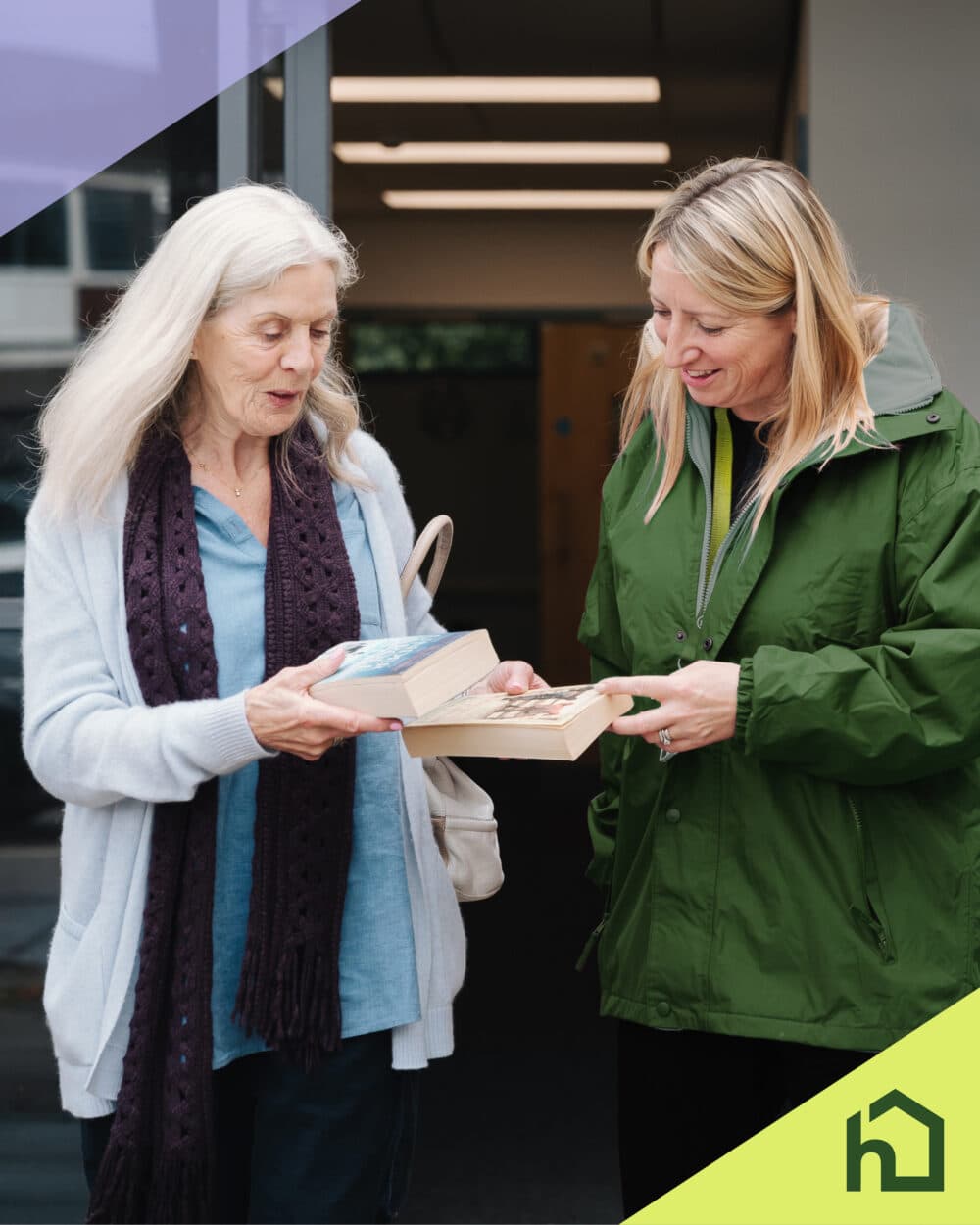 Two women smiling and exchanging books outside a building, with a green house logo in the corner. - Home Instead