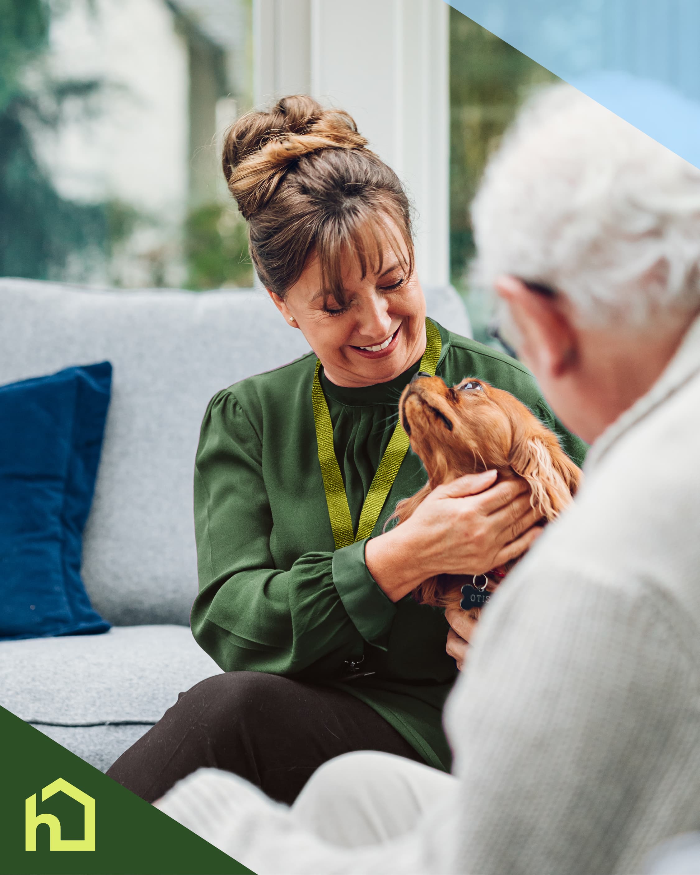 A woman smiles while petting a dog, sitting beside an older man on a couch. - Home Instead