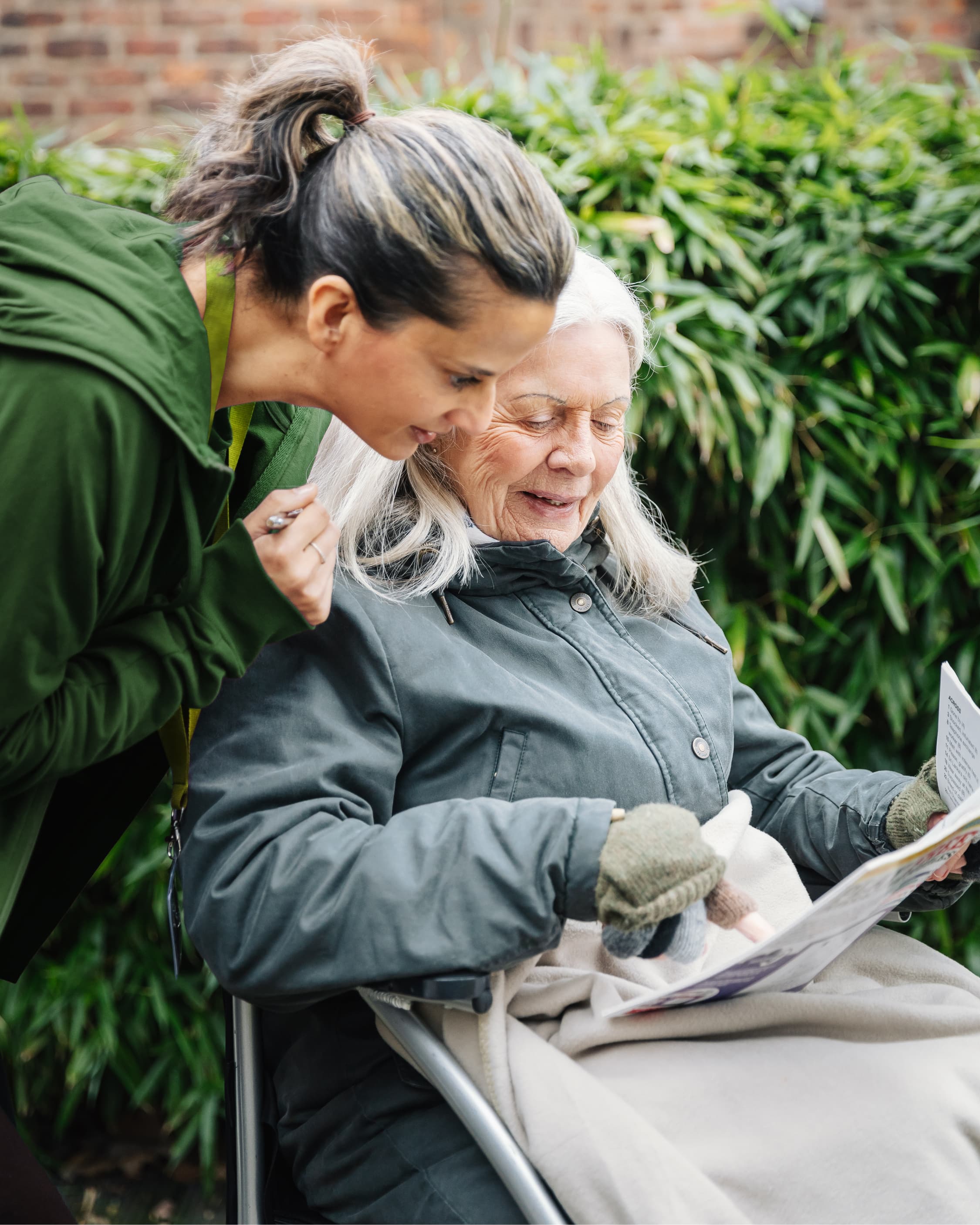 An older woman in a coat reads outdoors with a younger woman leaning in beside her, both smiling. - Home Instead