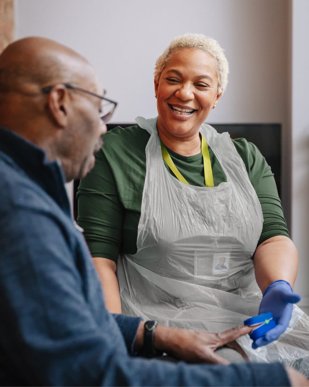 A smiling healthcare worker wearing gloves assists an older man with a medical test at home. - Home Instead