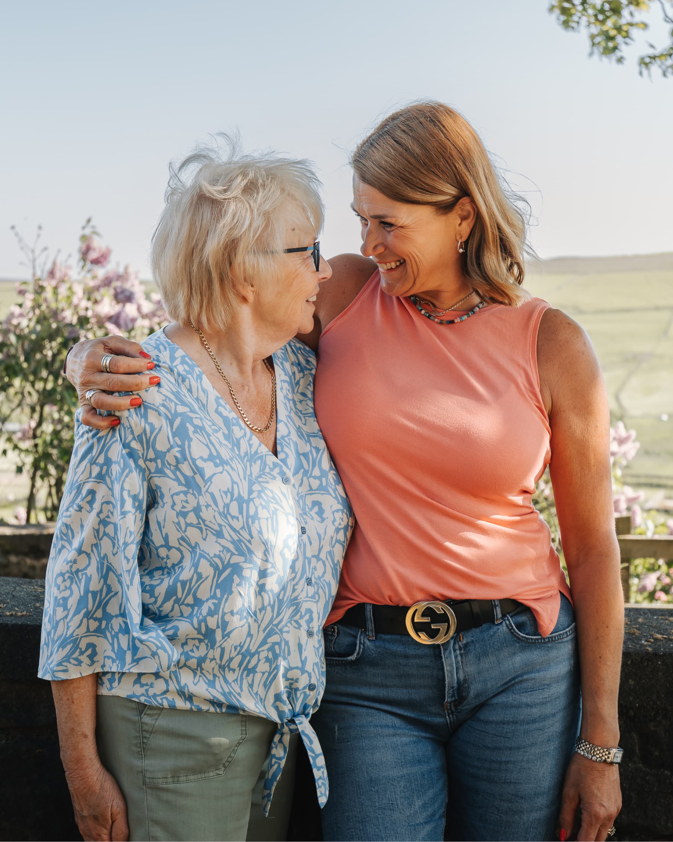 Two women smiling and embracing outdoors with green fields and flowers in the background. - Home Instead