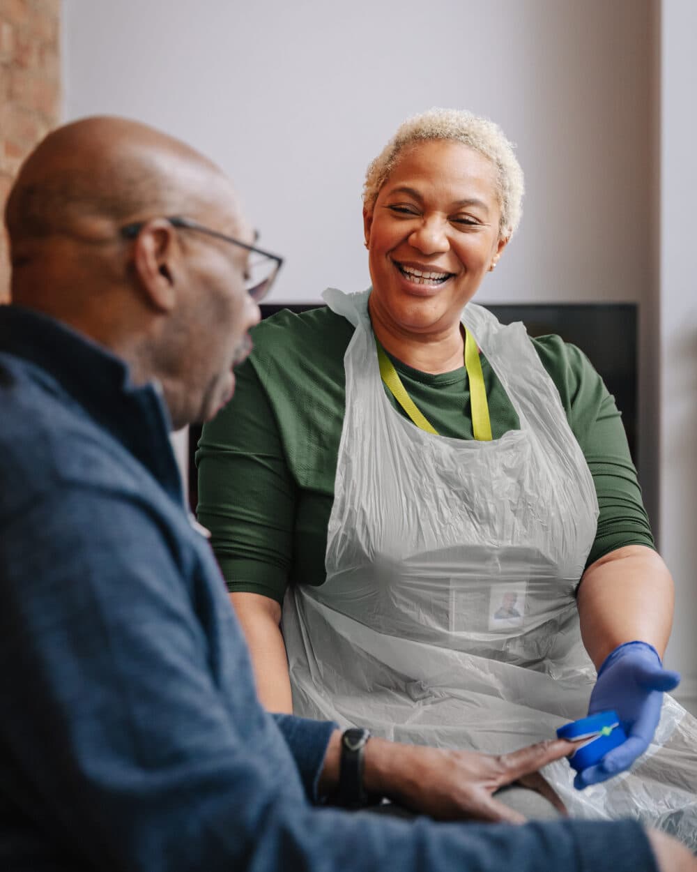 A smiling caregiver in gloves talks to an older man while checking his hand. - Home Instead
