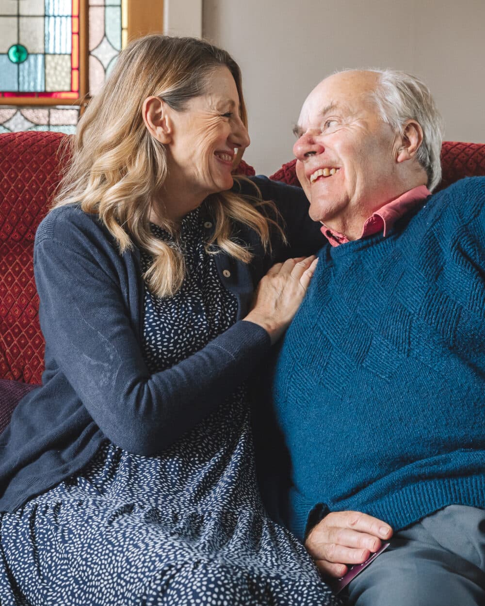 A woman and older man sit close together on a couch, smiling warmly at each other. - Home Instead