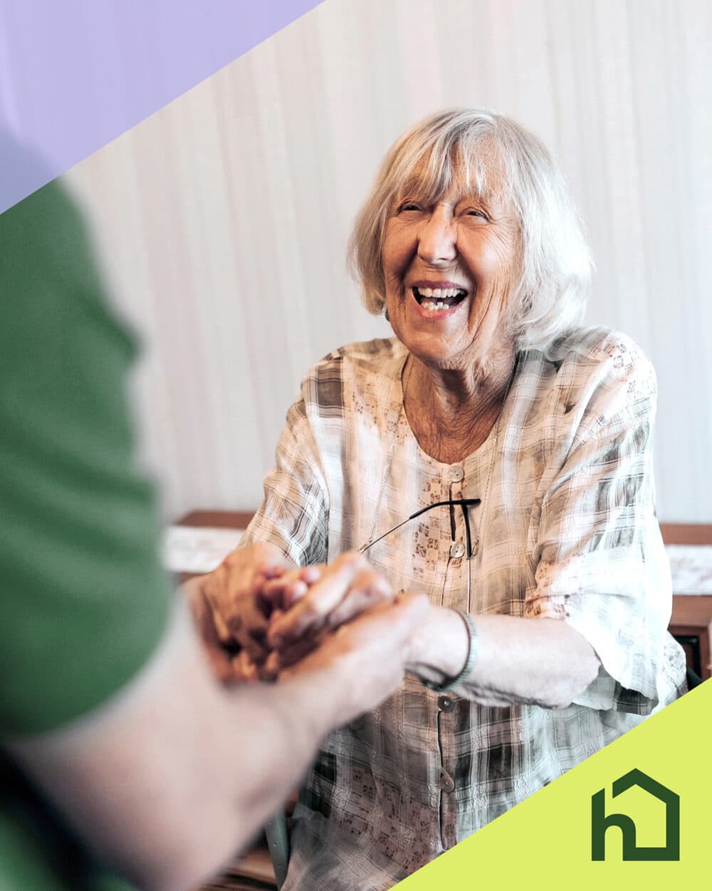 Smiling elderly woman holding hands with another person, sitting indoors; house logo in the corner. - Home Instead