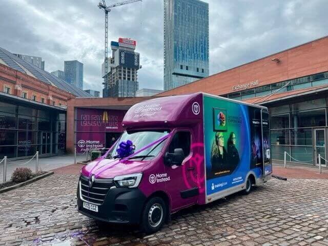 A colorful branded van is parked on a cobblestone path outside modern city buildings under a cloudy sky. - Home Instead