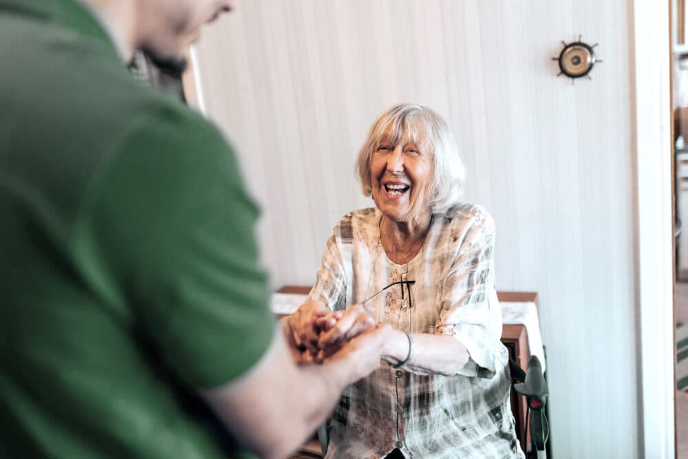 Smiling elderly woman sits and holds hands with a man in a green shirt, sharing a joyful moment indoors. - Home Instead