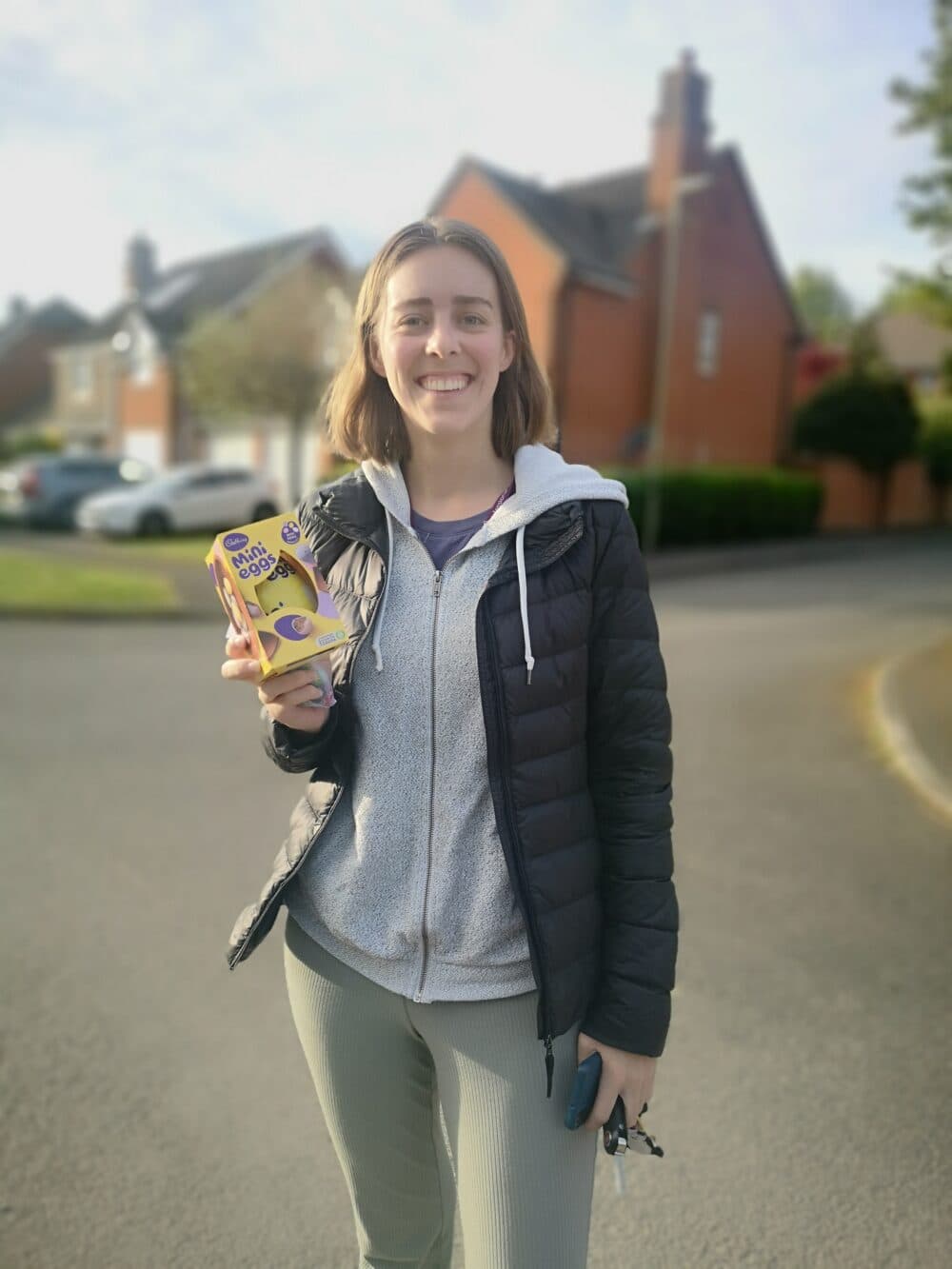 A smiling woman stands outdoors holding a Cadbury Easter egg box on a suburban street. - Home Instead
