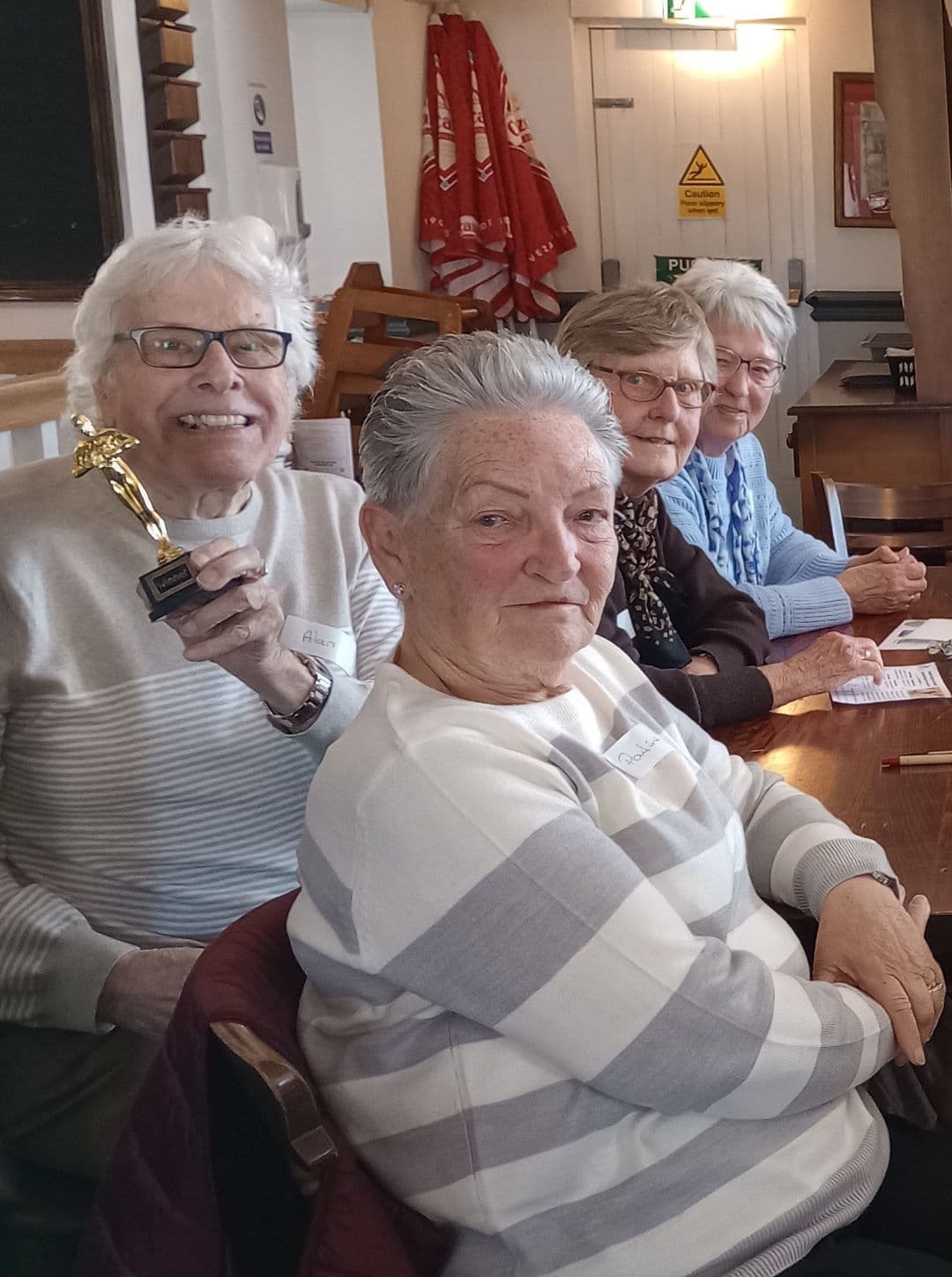 Four elderly women sit at a table; one smiles and holds a small gold trophy. - Home Instead