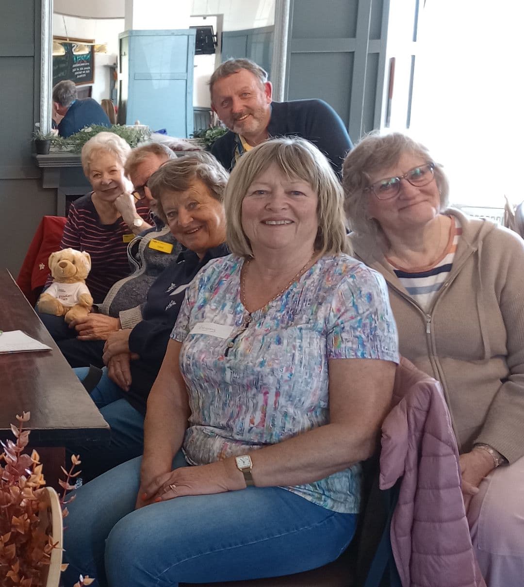 Six smiling older adults sit together indoors, one holding a teddy bear, posing for a group photo. - Home Instead