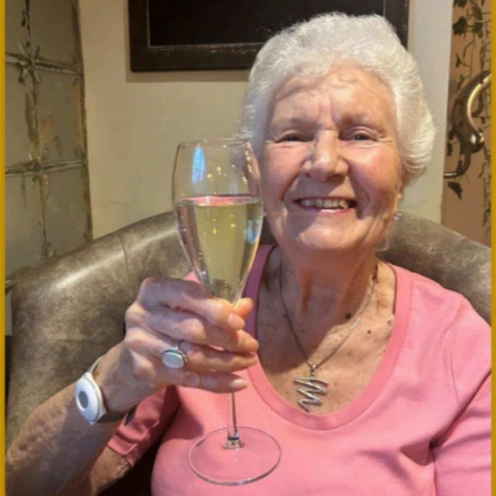 Smiling elderly woman in a pink shirt raises a glass of champagne while sitting in a chair. - Home Instead