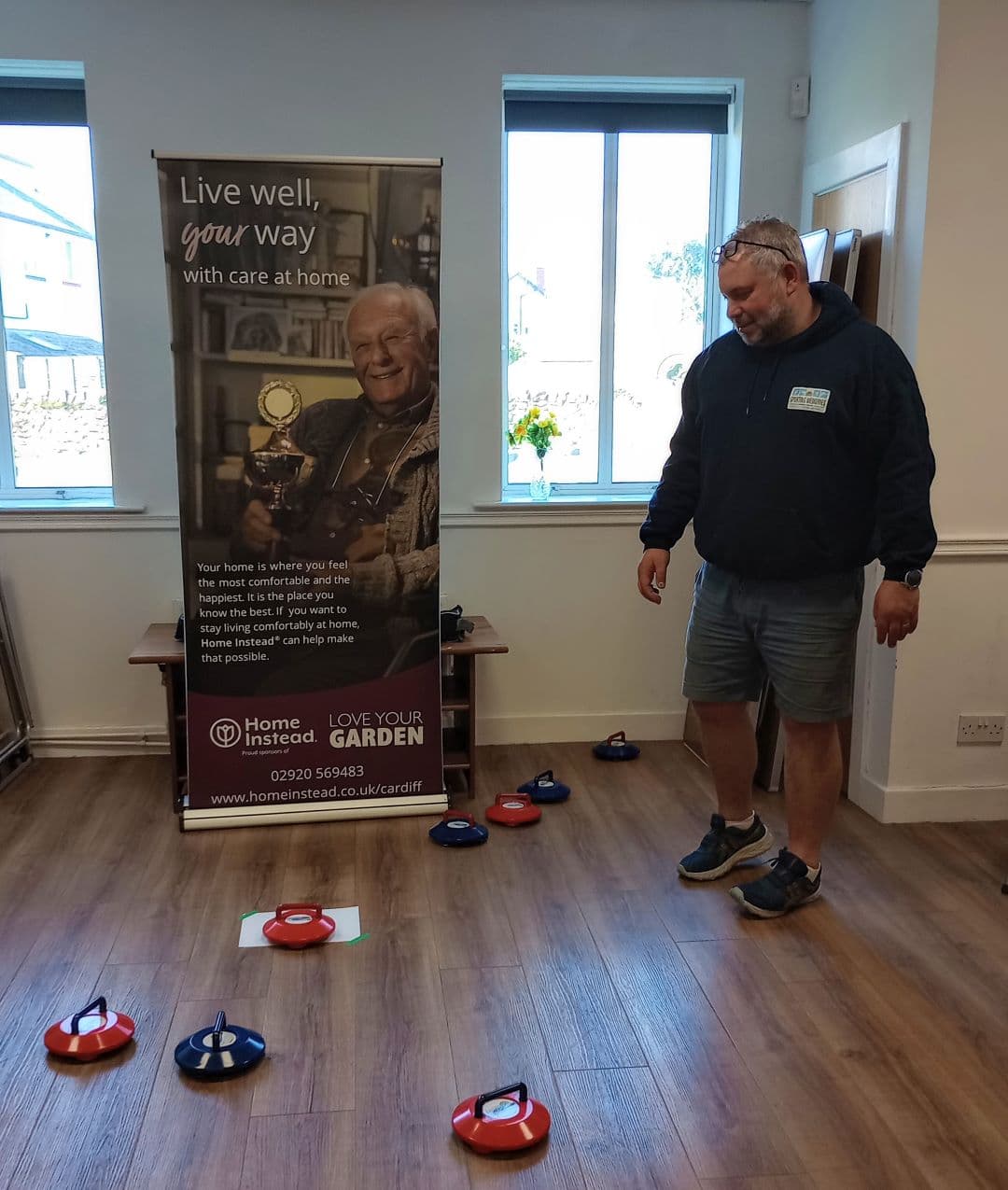 A man stands near curling stones indoors, next to a Home Instead care service banner and a window. - Home Instead