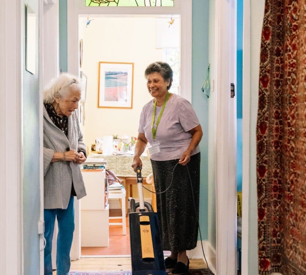 An older female adult chatting with her female carer with black hair and vacuuming the floor of the house