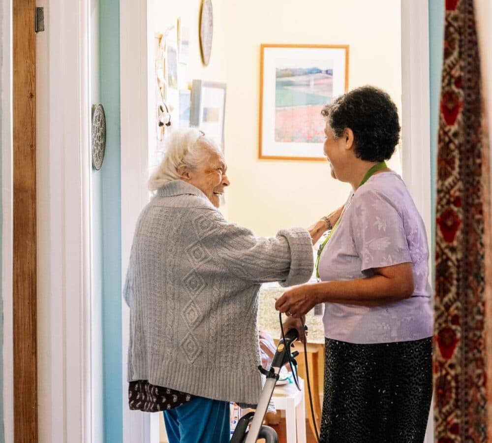 An older female adult with grey hair smiling while going inside the bathroom with the help of her female carer with black hair and holding a vacuum cleaner inside the house