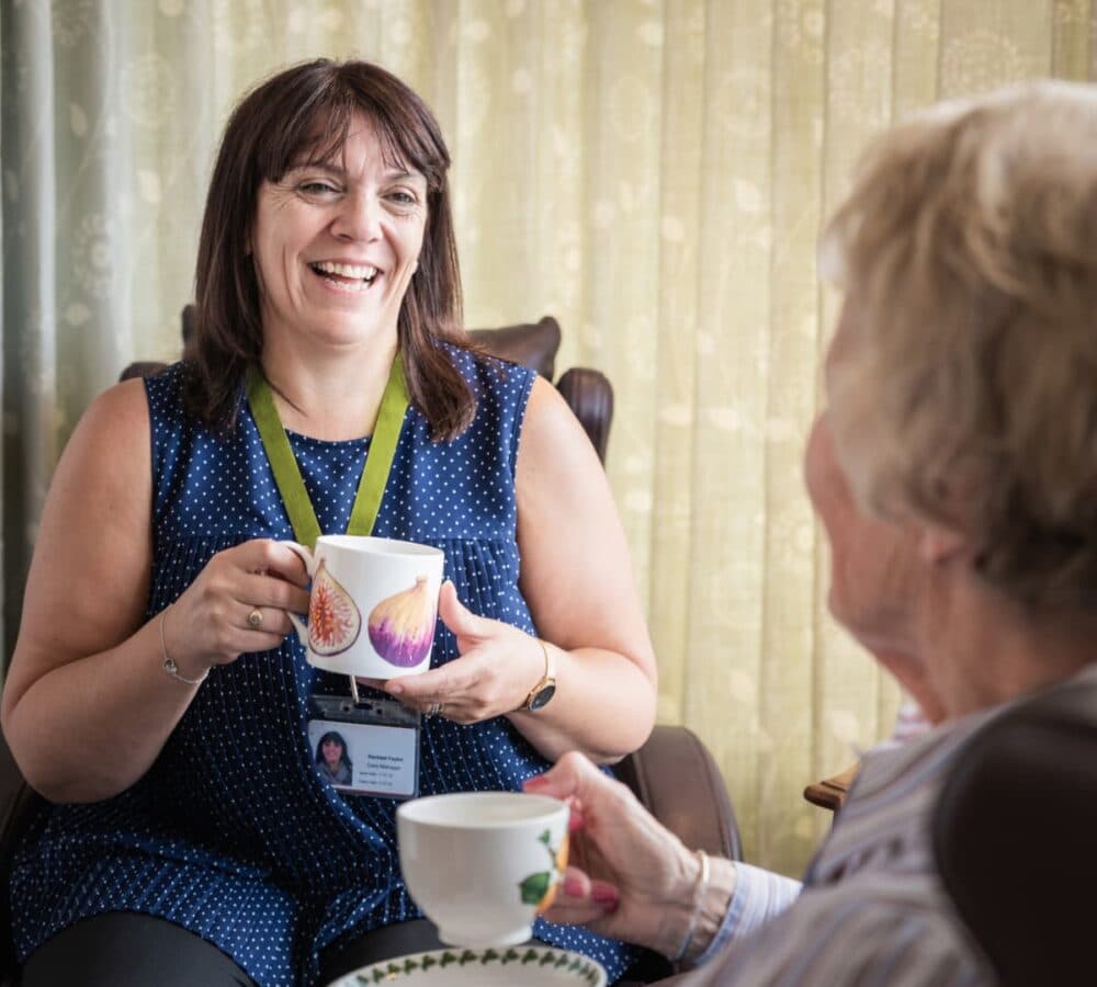 A woman happy and smiling while sitting and holding a cup of coffee and while chatting with an older female adult