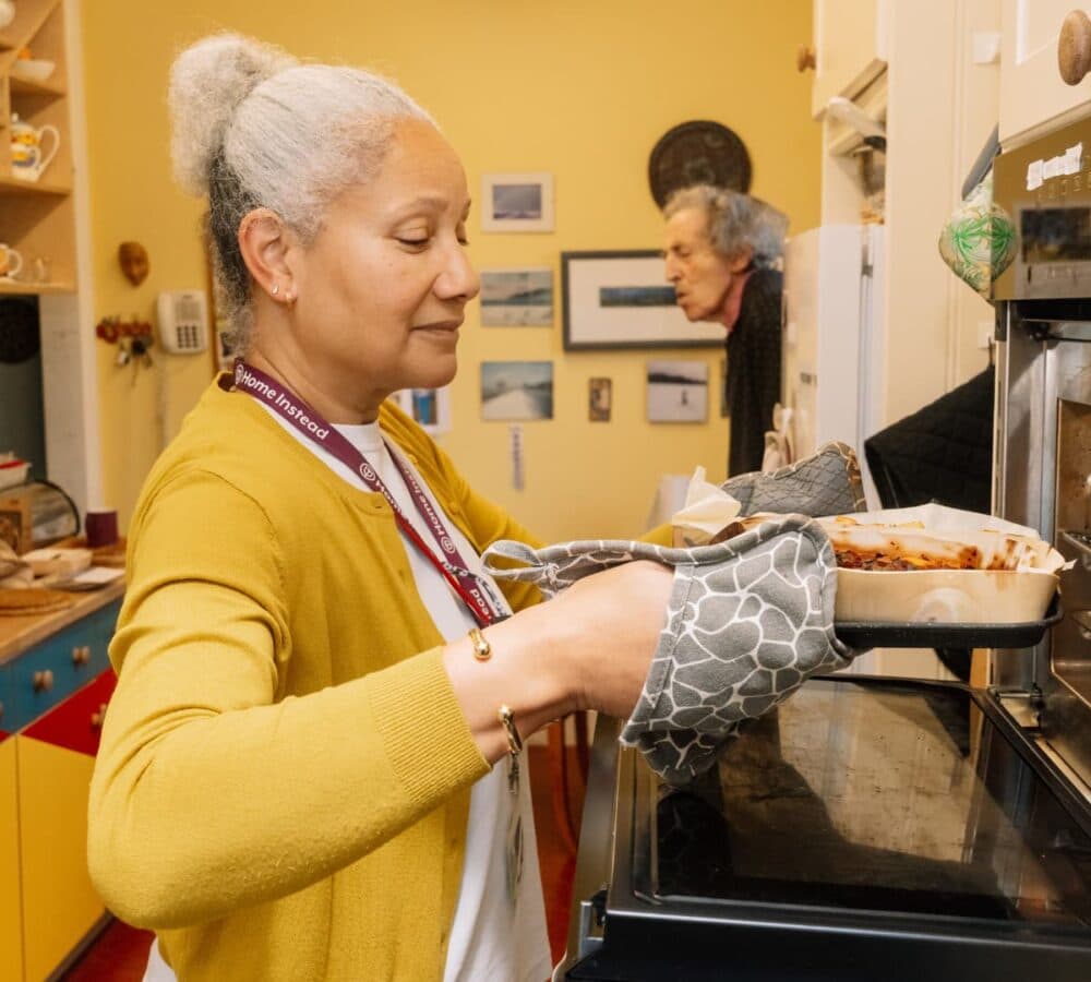 A woman with grey hair wearing yellow sweater and putting a pasta inside the oven