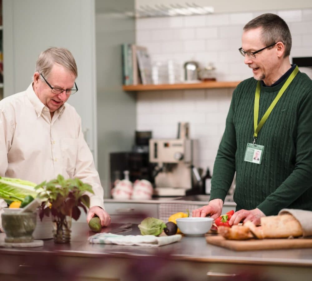A male carer wearing green and cutting vegetables with an older male adult inside the kitchen