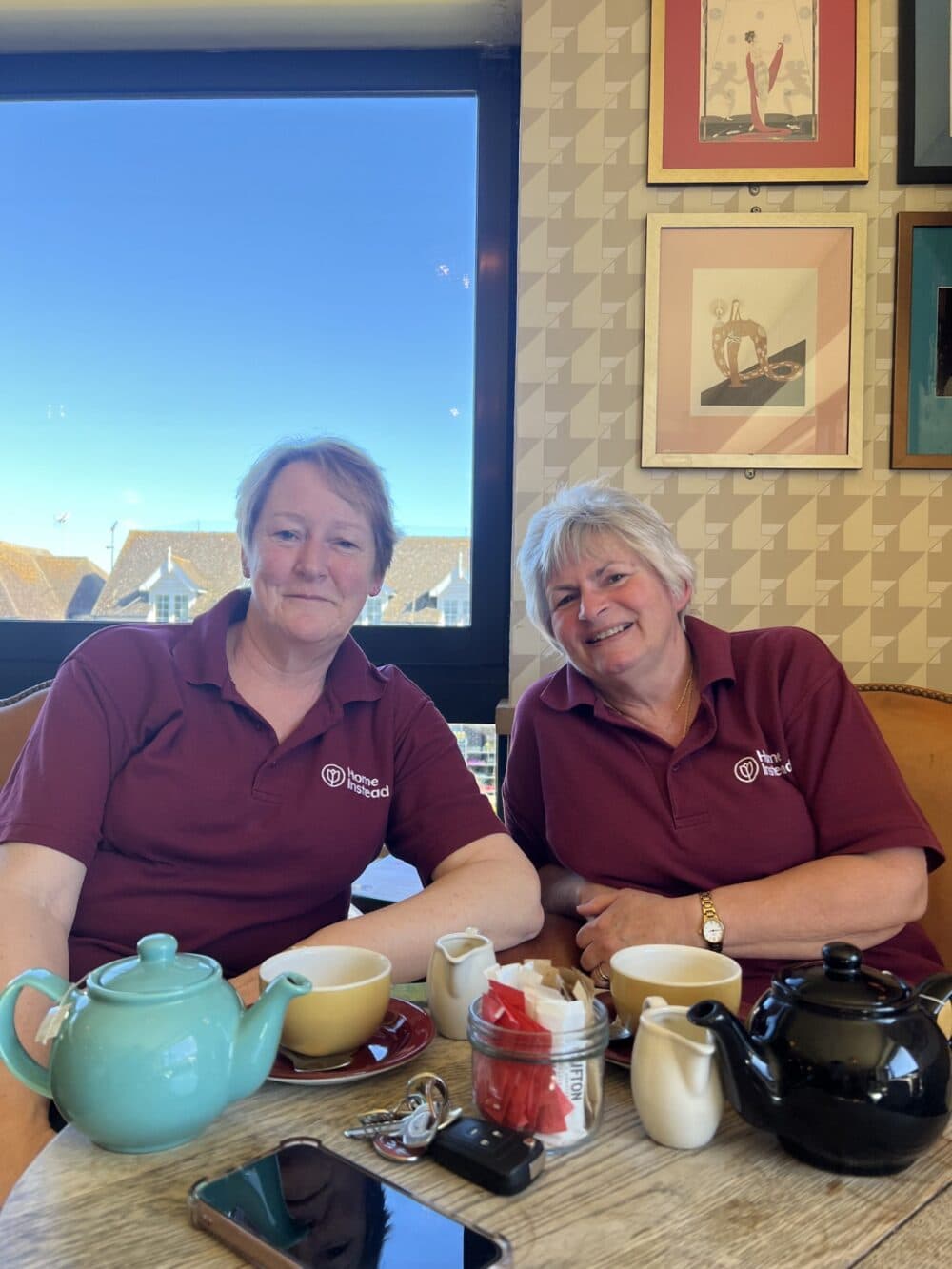 Two women in matching maroon shirts enjoying tea together at a café table, smiling at the camera. - Home Instead