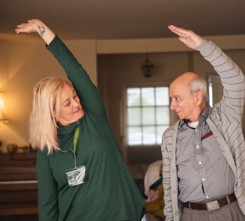 An older male adult exercising with his younger female carer wearing green and with long blonde hair inside the home