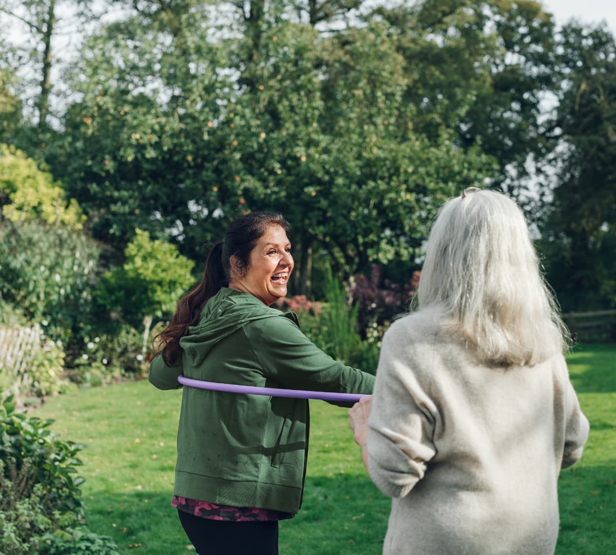 An older woman with grey hair and with her younger female carer wearing green and using the hula hop smiling while outdoors