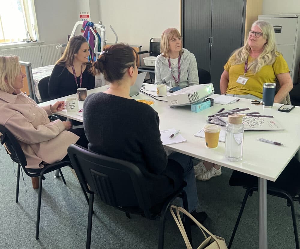 Five women sit around a table having a discussion with papers, drinks, and notebooks in front of them. - Home Instead