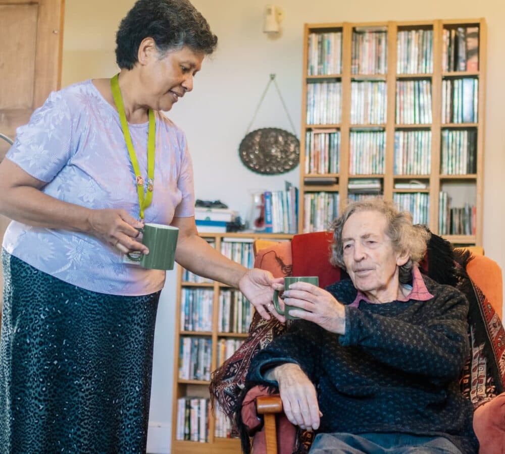 A woman giving a cup of coffee to an older male adult sitting on a couch inside the home