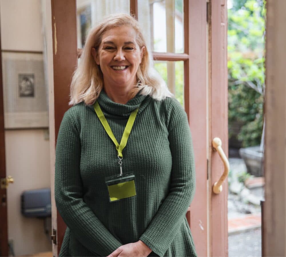 A woman with blonde hair and wearing green smiling near the door and inside the home