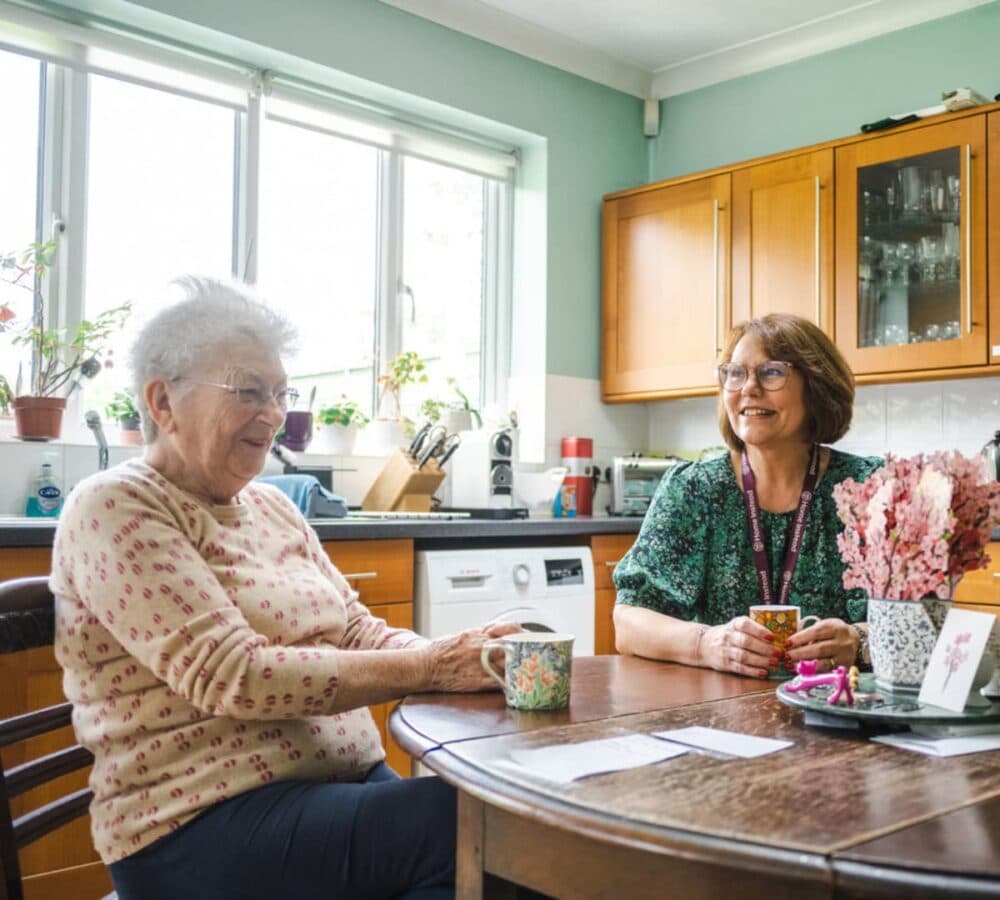 An older female adult with her younger female carer having coffee and both happy and smiling inside the kitchen