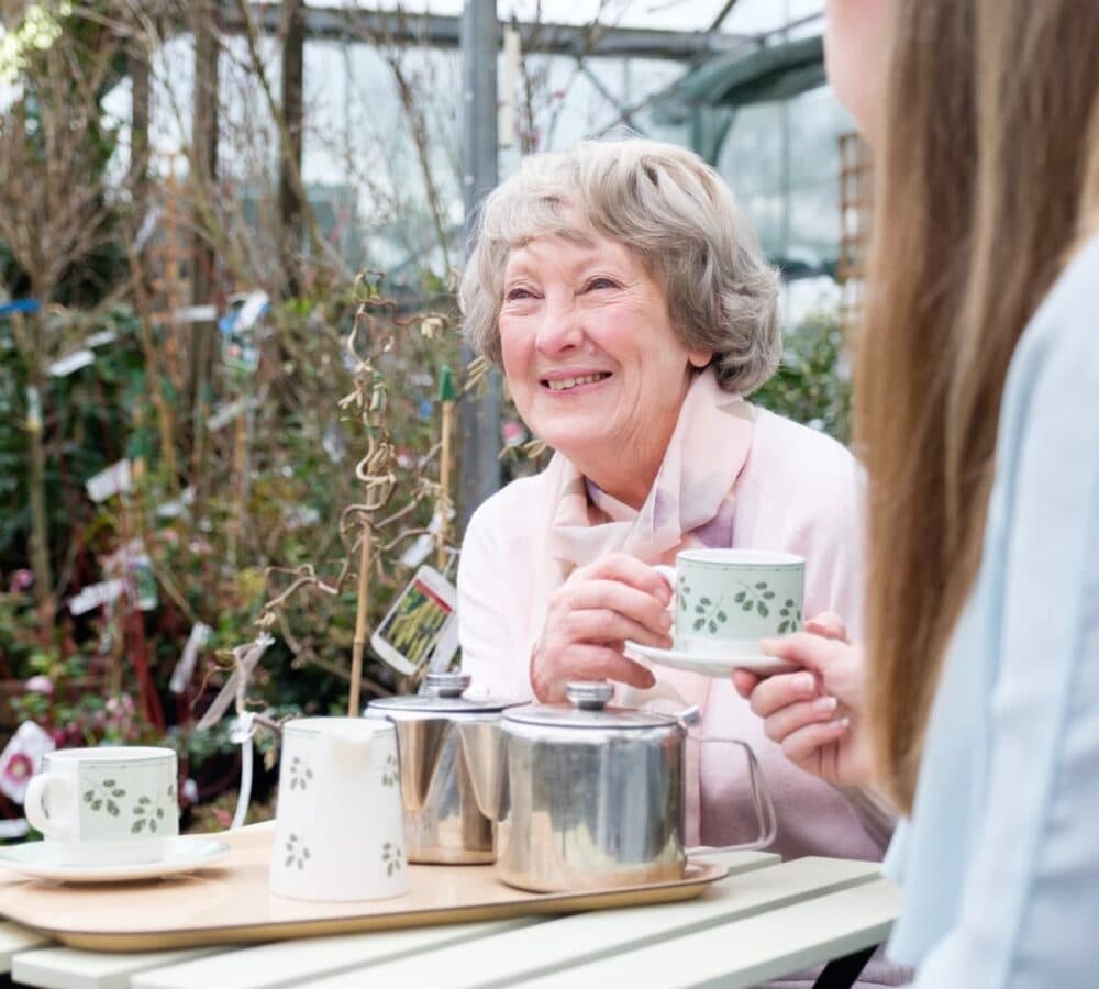 An older female adult with grey hair and wearing pink happy and smiling while having a cup of tea outdoors