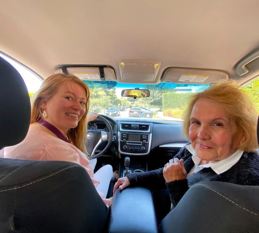 Two women smiling in the front seats of a car, looking back toward the camera. - Home Instead