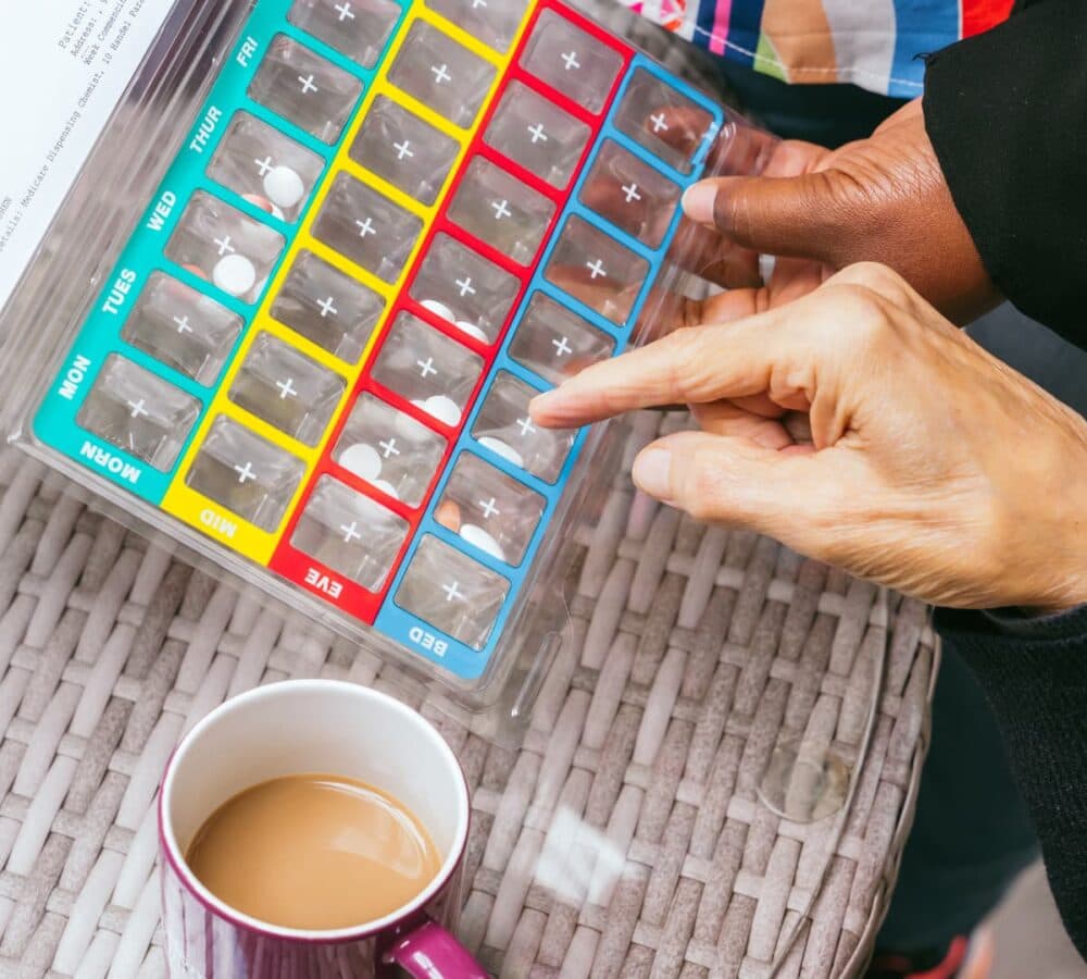 Medicine organizer in different colours with a cup of coffee on the side and fingers pointing aand holding it