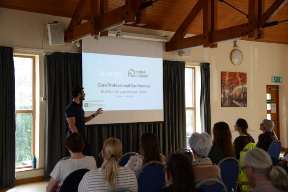 A man presents "Care Professional Conference" to an audience in a bright room with a projector screen. - Home Instead