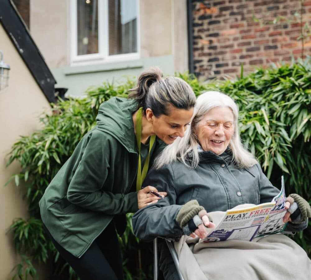 Older woman with white hair sitting on a wheelchair while smiling and looking at a crossword with her younger female carer outside the house
