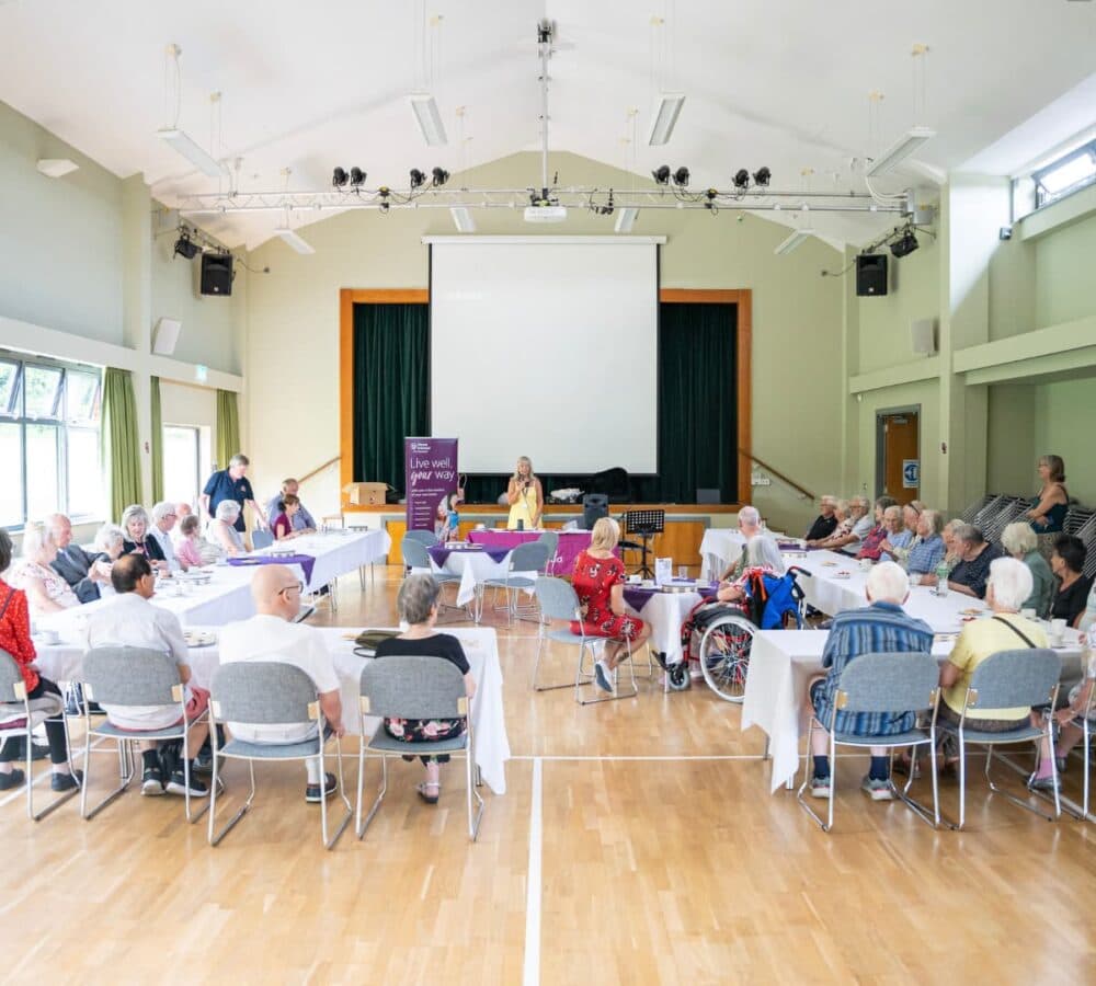 People inside the hall with a stage and a woman wearing yellow speaking in front