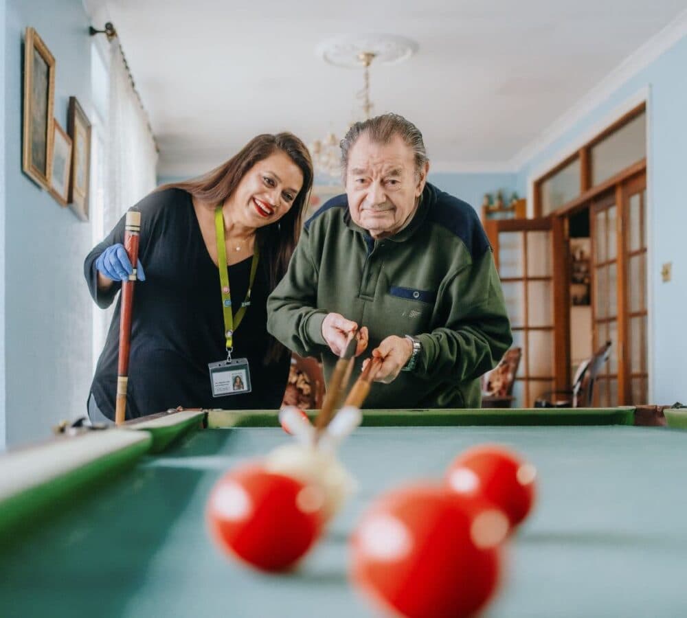 An older man with grey hair happily playing billiards with his younger female carer with long black hair and wearing gloves inside the house