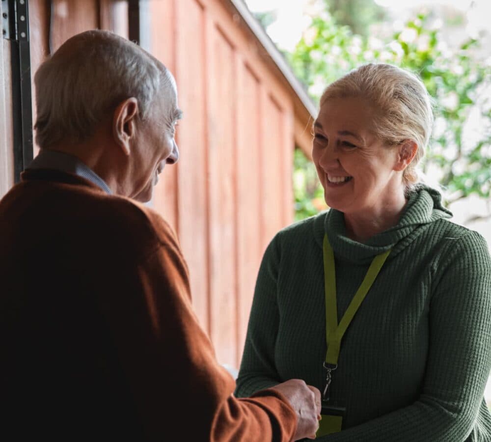 An older male adult with grey hair at the door welcoming his younger female carer wearing green both happy and smiling