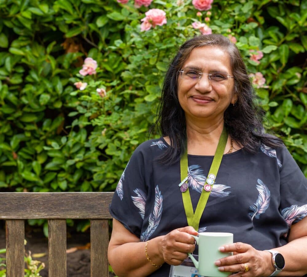 Smiling woman with glasses holding a mug, sitting on a bench in a garden with pink flowers. - Home Instead