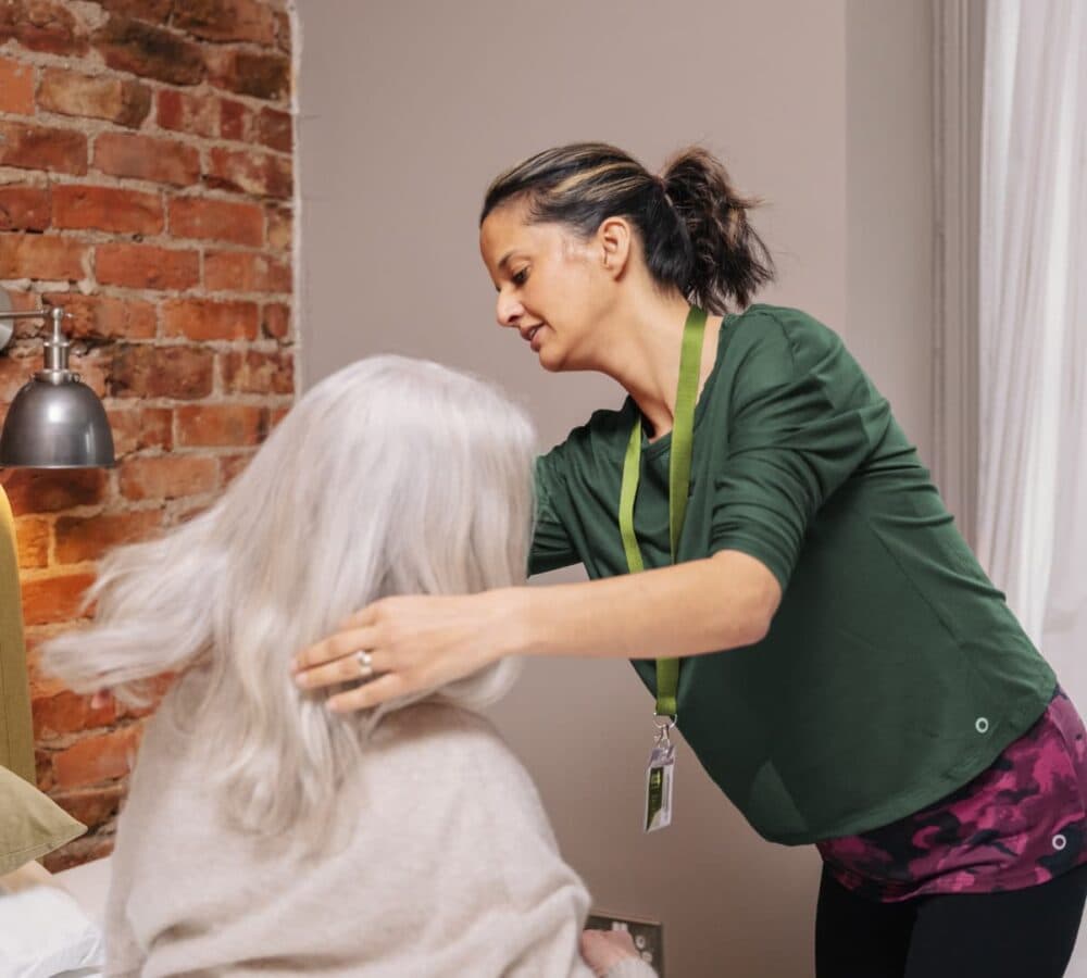 An older female woman sitting on her bed with long white hair and with her younger female carer fixing her hair while standing inside the room