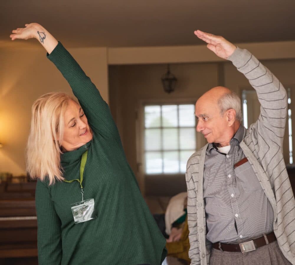 An older man with grey hair exercising with his younger female carer inside the house