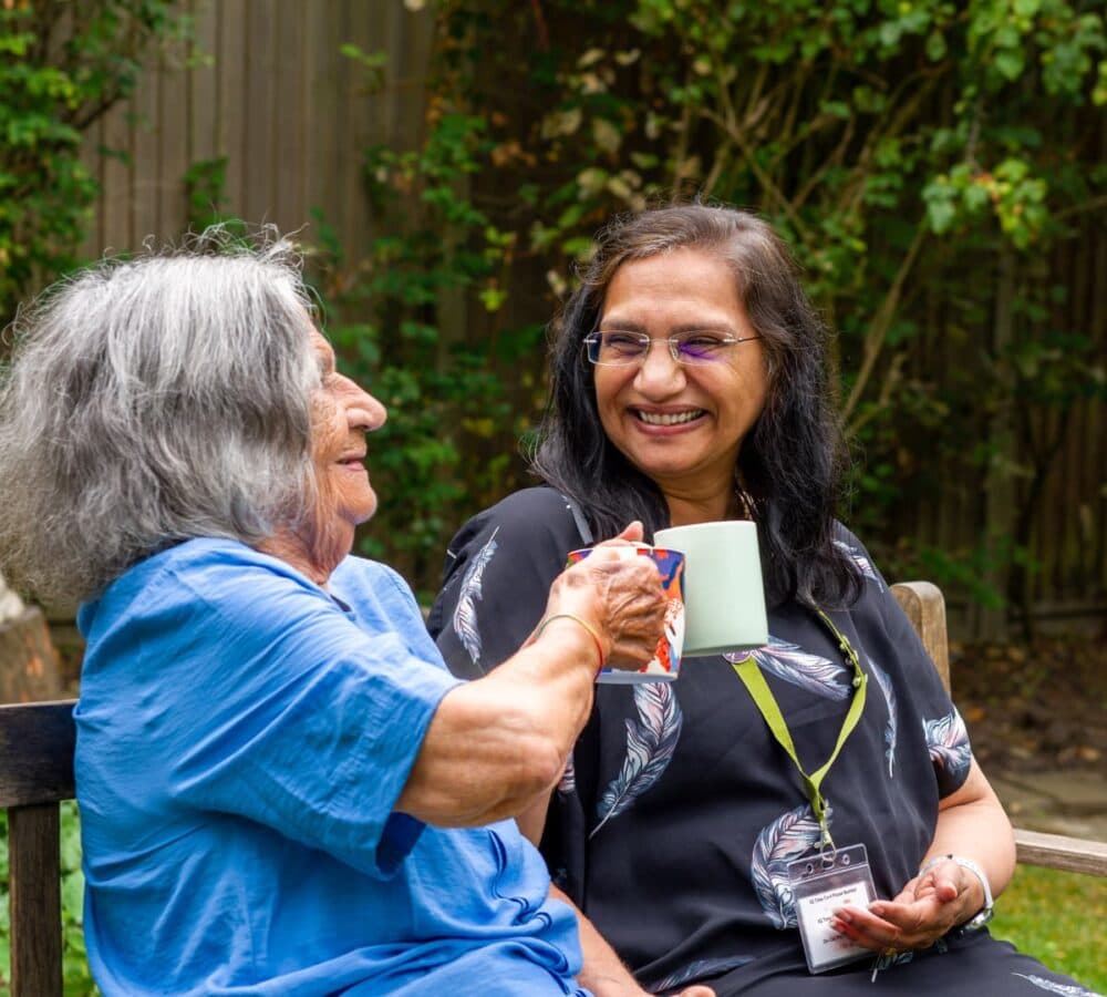 An older woman with grey hair and wearing blue sitting on a bench while toasting a cup of coffee with her younger female carer with long black hair and wearing a black top both happy and smiling
